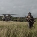 A fully equipped military soldier in camouflage gear walks through a grassy field, moving away from a large tandem-rotor helicopter in the background. The setting appears to be a tropical or subtropical environment with dense tree lines and overcast skies. The scene suggests a recent helicopter insertion or extraction operation.