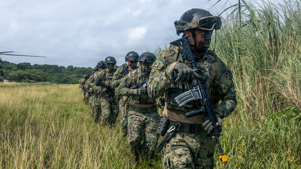 A column of military personnel in camouflage uniforms and tactical helmets moves through a grassy field in single file. The soldiers are armed and equipped with full combat gear, appearing to conduct a field exercise or tactical movement. A helicopter is partially visible in the background left, suggesting an air insertion or extraction scenario.