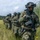 PANAMAX-26: V1/6, Partner forces conduct simulated air raid A column of military personnel in camouflage uniforms and tactical helmets moves through a grassy field in single file. The soldiers are armed and equipped with full combat gear, appearing to conduct a field exercise or tactical movement. A helicopter is partially visible in the background left, suggesting an air insertion or extraction scenario.