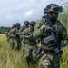 A column of military personnel in camouflage uniforms and tactical helmets moves through a grassy field in single file. The soldiers are armed and equipped with full combat gear, appearing to conduct a field exercise or tactical movement. A helicopter is partially visible in the background left, suggesting an air insertion or extraction scenario.