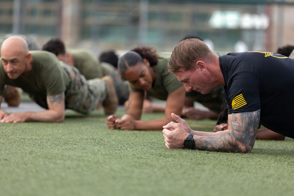 A group of military personnel are performing plank exercises on an outdoor artificial turf field. The individuals are wearing a mix of military uniforms, including olive/camouflage PT gear and a black uniform with gold insignia. The foreground subject is a male with visible tattoos and a sports watch, holding a forearm plank position.