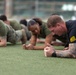 A group of military personnel are performing plank exercises on an outdoor artificial turf field. The individuals are wearing a mix of military uniforms, including olive/camouflage PT gear and a black uniform with gold insignia. The foreground subject is a male with visible tattoos and a sports watch, holding a forearm plank position.