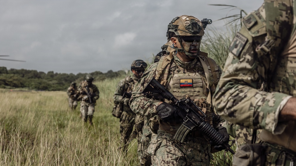 A group of heavily equipped military personnel are moving through an open grassy field in tactical formation. The lead soldier prominently displays an Ecuadorian flag patch on their plate carrier vest and wears full combat gear including a ballistic helmet with night vision mount, tactical eyewear, and face paint. The scene appears to be a military operation or training exercise in a rural environment.