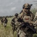 A group of heavily equipped military personnel are moving through an open grassy field in tactical formation. The lead soldier prominently displays an Ecuadorian flag patch on their plate carrier vest and wears full combat gear including a ballistic helmet with night vision mount, tactical eyewear, and face paint. The scene appears to be a military operation or training exercise in a rural environment.