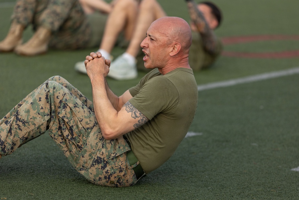 A bald male in U.S. Marine Corps MARPAT camouflage trousers and an olive drab t-shirt performs crunches/sit-ups on an athletic turf field. His hands are clasped together at chest level and his expression shows physical exertion. Additional personnel in similar uniforms are visible but out of focus in the background.