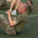 A bald male in U.S. Marine Corps MARPAT camouflage trousers and an olive drab t-shirt performs crunches/sit-ups on an athletic turf field. His hands are clasped together at chest level and his expression shows physical exertion. Additional personnel in similar uniforms are visible but out of focus in the background.