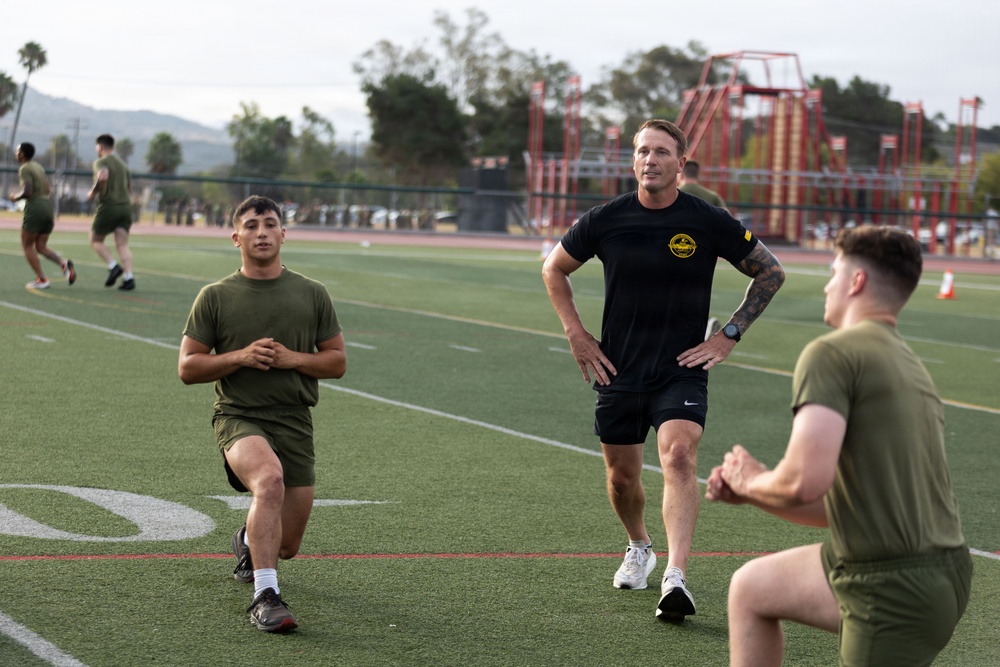 A physical training session is taking place on an outdoor turf athletic field, likely at a military installation. A man in a black t-shirt with a circular logo patch appears to be an instructor or supervisor observing two younger individuals performing walking lunges. Additional personnel in olive drab PT uniforms are visible running in the background.