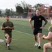 A physical training session is taking place on an outdoor turf athletic field, likely at a military installation. A man in a black t-shirt with a circular logo patch appears to be an instructor or supervisor observing two younger individuals performing walking lunges. Additional personnel in olive drab PT uniforms are visible running in the background.
