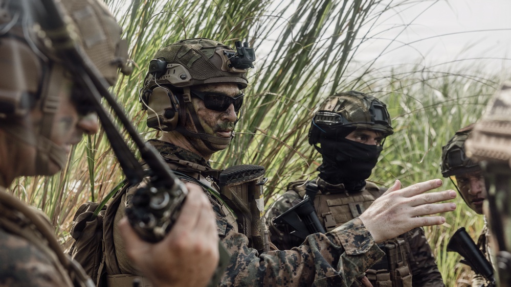 The image shows a group of military personnel in full tactical gear gathered in a dense vegetation area, appearing to conduct a briefing or coordination. At least three soldiers are visible, wearing camouflage uniforms, tactical helmets, and body armor. One soldier is holding a radio while another appears to be gesturing, suggesting active communication or mission planning.