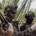 The image shows a group of military personnel in full tactical gear gathered in a dense vegetation area, appearing to conduct a briefing or coordination. At least three soldiers are visible, wearing camouflage uniforms, tactical helmets, and body armor. One soldier is holding a radio while another appears to be gesturing, suggesting active communication or mission planning.