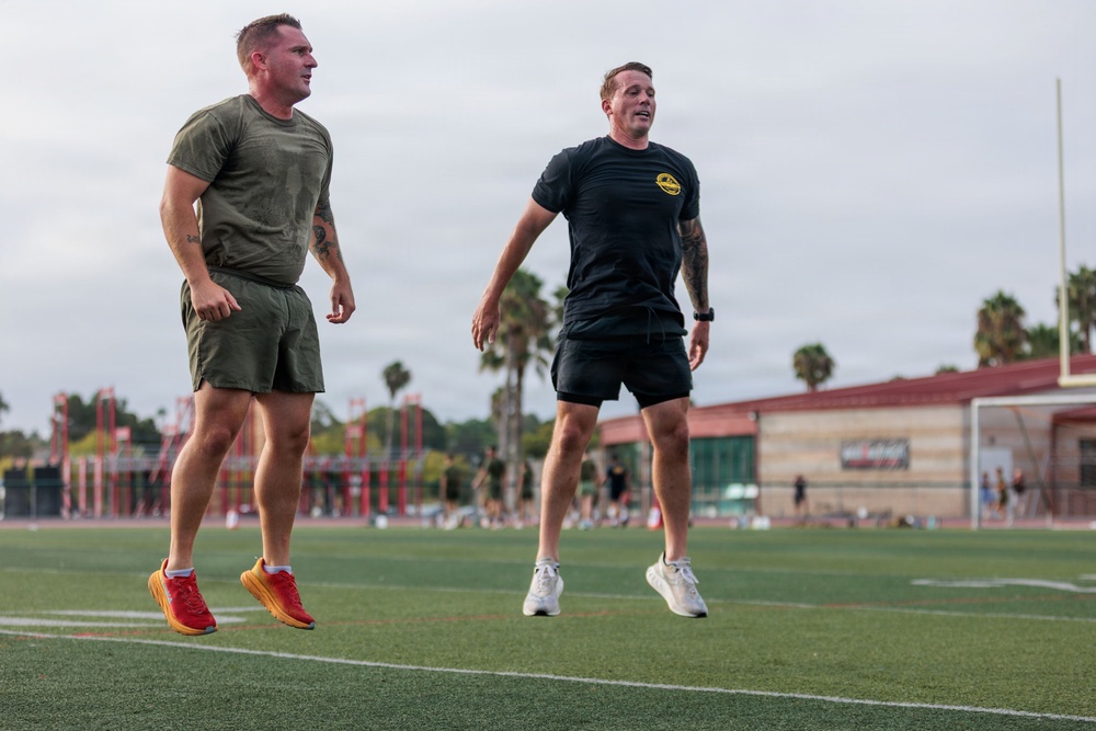 Two athletic men are exercising on a green turf athletic field, both captured mid-air suggesting jumping or running drills. They are dressed in athletic wear and appear to be engaged in an outdoor workout session. The background shows palm trees, a sports facility building, and additional people on the track in the distance.