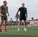 Two athletic men are exercising on a green turf athletic field, both captured mid-air suggesting jumping or running drills. They are dressed in athletic wear and appear to be engaged in an outdoor workout session. The background shows palm trees, a sports facility building, and additional people on the track in the distance.