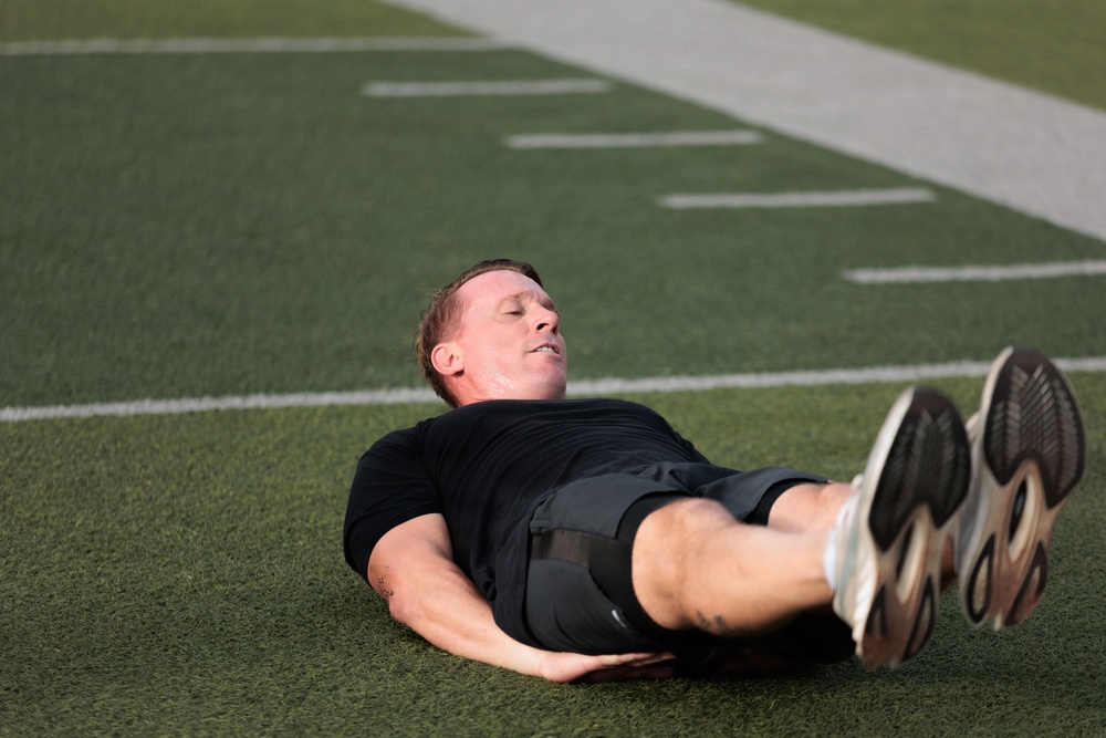 A man wearing black athletic clothing is lying on his back on a green artificial turf football field. His eyes are closed and legs are raised slightly, suggesting he is resting or recovering after physical exertion. White yard line markings are visible on the turf in the background.