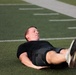A man wearing black athletic clothing is lying on his back on a green artificial turf football field. His eyes are closed and legs are raised slightly, suggesting he is resting or recovering after physical exertion. White yard line markings are visible on the turf in the background.