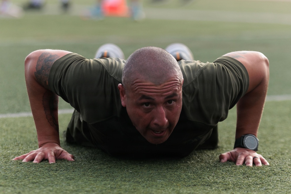A bald man wearing an olive/dark green athletic shirt is performing push-ups on a green turf field. He is in the down position of the push-up, with his chest near the ground and his face looking directly at the camera. He has visible tattoos on his arms and is wearing a dark sports watch on his right wrist.