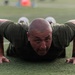 A bald man wearing an olive/dark green athletic shirt is performing push-ups on a green turf field. He is in the down position of the push-up, with his chest near the ground and his face looking directly at the camera. He has visible tattoos on his arms and is wearing a dark sports watch on his right wrist.