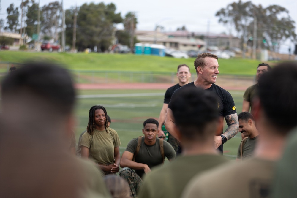 A man in a black athletic shirt with visible tattoos on his arm stands and speaks to a group of individuals wearing olive drab military-style physical training uniforms, who are seated on a field. The setting appears to be a military installation or base, with a running track, green grass, and institutional buildings visible in the background. The atmosphere appears relaxed and engaged, suggesting a post-exercise briefing or motivational talk.
