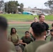 A man in a black athletic shirt with visible tattoos on his arm stands and speaks to a group of individuals wearing olive drab military-style physical training uniforms, who are seated on a field. The setting appears to be a military installation or base, with a running track, green grass, and institutional buildings visible in the background. The atmosphere appears relaxed and engaged, suggesting a post-exercise briefing or motivational talk.