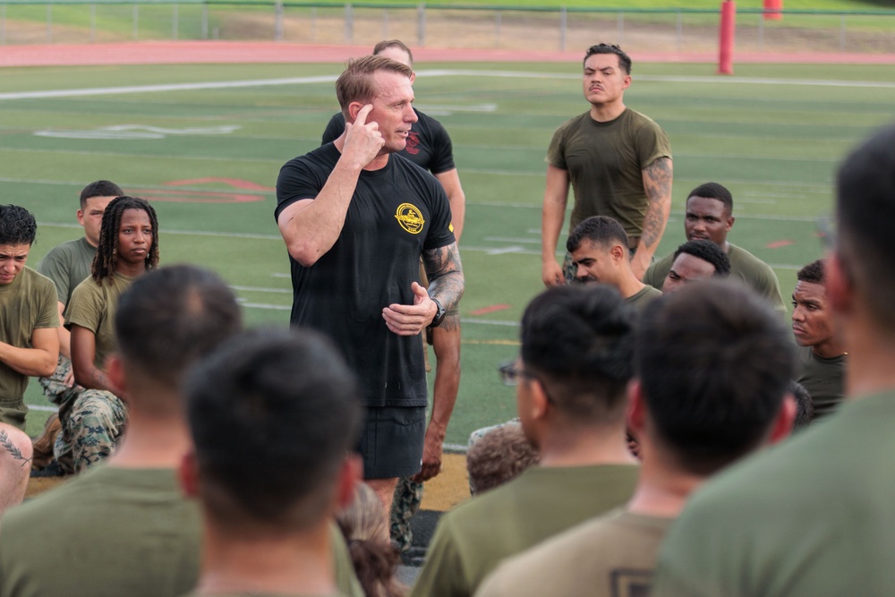 A man in a black t-shirt with a yellow emblem is addressing a large group of military personnel in olive drab uniforms seated on a sports field. He appears to be an instructor or leader conducting a briefing or training session. The setting is an outdoor athletic field with visible yard lines and a running track in the background.