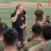 A man in a black t-shirt with a yellow emblem is addressing a large group of military personnel in olive drab uniforms seated on a sports field. He appears to be an instructor or leader conducting a briefing or training session. The setting is an outdoor athletic field with visible yard lines and a running track in the background.