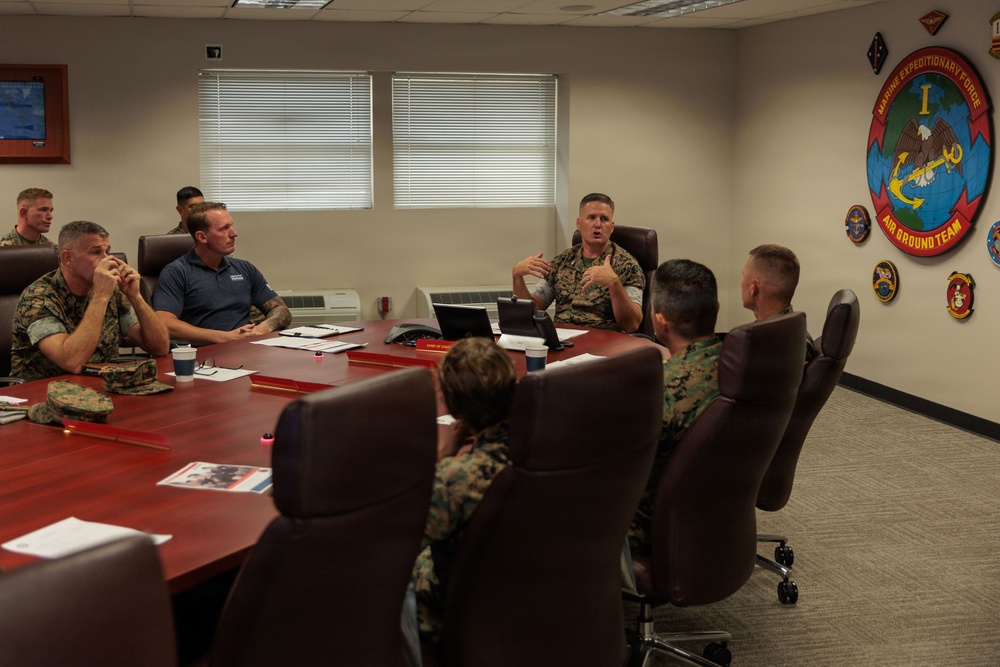 A military briefing or meeting is taking place in a formal conference room at what appears to be a I Marine Expeditionary Force (I MEF) facility. Several personnel in USMC MARPAT camouflage uniforms are seated around a large wooden conference table, along with at least one civilian. A senior Marine officer appears to be leading the discussion, gesturing while speaking.