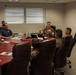 A military briefing or meeting is taking place in a formal conference room at what appears to be a I Marine Expeditionary Force (I MEF) facility. Several personnel in USMC MARPAT camouflage uniforms are seated around a large wooden conference table, along with at least one civilian. A senior Marine officer appears to be leading the discussion, gesturing while speaking.