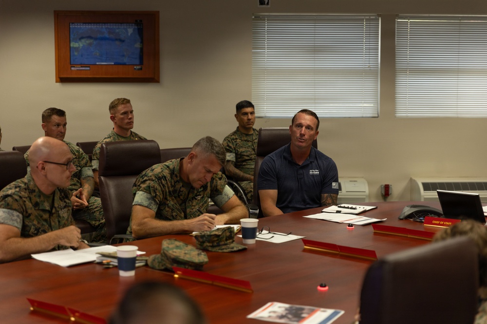 A military briefing or meeting is taking place in a conference room, with several U.S. Marines in camouflage uniforms seated around a large wooden table. A civilian in a navy blue polo shirt with an American flag emblem appears to be speaking or presenting to the group. A monitor displaying a map is visible on the wall in the background.