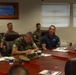 A military briefing or meeting is taking place in a conference room, with several U.S. Marines in camouflage uniforms seated around a large wooden table. A civilian in a navy blue polo shirt with an American flag emblem appears to be speaking or presenting to the group. A monitor displaying a map is visible on the wall in the background.