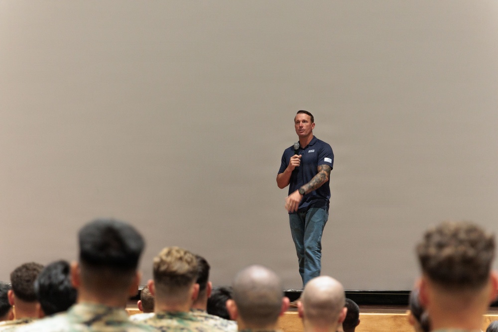 A man in a navy blue polo shirt and jeans stands on a stage holding a microphone, addressing a seated audience. The audience members, several wearing military camouflage uniforms, are viewed from behind. The setting appears to be an auditorium or theater-style venue with a large blank projection screen in the background.