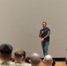 A man in a navy blue polo shirt and jeans stands on a stage holding a microphone, addressing a seated audience. The audience members, several wearing military camouflage uniforms, are viewed from behind. The setting appears to be an auditorium or theater-style venue with a large blank projection screen in the background.