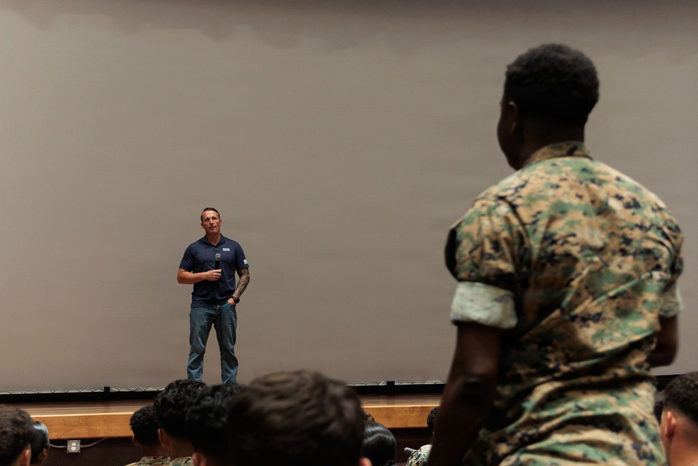 A civilian speaker in a navy blue polo shirt and jeans stands on a stage addressing an audience of military personnel in camouflage uniforms. The setting appears to be an auditorium or briefing room with a large projection screen behind the speaker. A Marine in MARPAT woodland camouflage is prominently visible in the foreground, appearing to stand or ask a question.