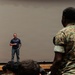 A civilian speaker in a navy blue polo shirt and jeans stands on a stage addressing an audience of military personnel in camouflage uniforms. The setting appears to be an auditorium or briefing room with a large projection screen behind the speaker. A Marine in MARPAT woodland camouflage is prominently visible in the foreground, appearing to stand or ask a question.