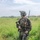 PANAMAX-26: V1/6, Partner forces conduct simulated air raid A fully equipped military service member stands in a tall grass field in what appears to be a tropical environment. The soldier is wearing digital camouflage uniform consistent with USMC MARPAT pattern, full tactical gear, and is carrying an assault rifle. A building with a red roof and palm trees are visible in the background.