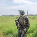 A fully equipped military service member stands in a tall grass field in what appears to be a tropical environment. The soldier is wearing digital camouflage uniform consistent with USMC MARPAT pattern, full tactical gear, and is carrying an assault rifle. A building with a red roof and palm trees are visible in the background.