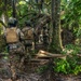 A group of military personnel in full tactical gear are moving through dense jungle terrain in a single-file patrol formation. The soldiers are equipped with helmets, body armor, and tactical vests, suggesting a training exercise or operational patrol. The environment features thick tropical vegetation with fallen logs and palm fronds on the forest floor.