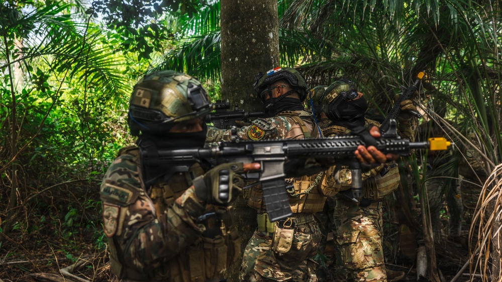 The image shows multiple armed military or special operations personnel in full tactical gear operating in a dense jungle or tropical forest environment. The soldiers are wearing camouflage uniforms, tactical helmets, and face coverings, and are carrying assault rifles in a ready position. Visible patches suggest multinational participation, possibly a joint training exercise.