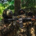 The image shows multiple armed military or special operations personnel in full tactical gear operating in a dense jungle or tropical forest environment. The soldiers are wearing camouflage uniforms, tactical helmets, and face coverings, and are carrying assault rifles in a ready position. Visible patches suggest multinational participation, possibly a joint training exercise.