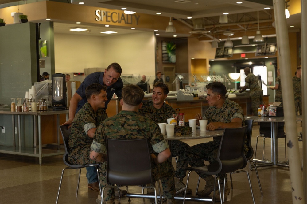 A group of military personnel in camouflage uniforms are seated at a round table in what appears to be a military dining facility (DFAC/mess hall). A civilian man in a blue shirt and jeans is leaning over the table, engaging in conversation with the service members. The setting is a modern, well-lit cafeteria-style facility with a 'Specialty' food station visible in the background.