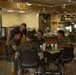 A group of military personnel in camouflage uniforms are seated at a round table in what appears to be a military dining facility (DFAC/mess hall). A civilian man in a blue shirt and jeans is leaning over the table, engaging in conversation with the service members. The setting is a modern, well-lit cafeteria-style facility with a 'Specialty' food station visible in the background.