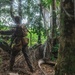 The image shows at least two individuals in military or tactical camouflage gear moving through a dense tropical jungle environment. The primary figure is visible from behind, carrying a backpack and equipment, appearing to navigate through fallen logs and thick vegetation. A second individual is partially visible behind a large tree trunk on the right side of the frame.