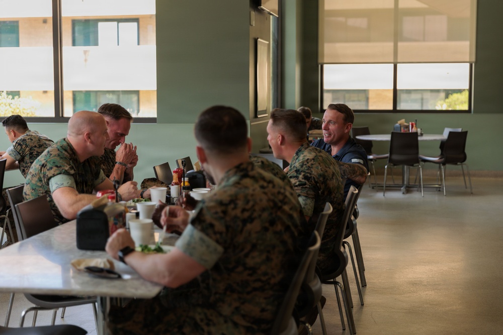 A group of military personnel in camouflage uniforms are seated together at a dining table in what appears to be a military mess hall or cafeteria. They are engaged in conversation during a meal, with one individual in civilian clothing (navy blue shirt) participating in the discussion. The setting is a modern, institutional dining facility with green walls, large windows, and standard cafeteria-style furniture.