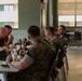 A group of military personnel in camouflage uniforms are seated together at a dining table in what appears to be a military mess hall or cafeteria. They are engaged in conversation during a meal, with one individual in civilian clothing (navy blue shirt) participating in the discussion. The setting is a modern, institutional dining facility with green walls, large windows, and standard cafeteria-style furniture.