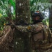 A uniformed soldier wearing Panamanian flag patch on helmet is positioned behind a tree in a tropical jungle environment, aiming a rifle. The soldier is equipped with full tactical gear including a plate carrier, backpack, and helmet with optics. The setting appears to be a training exercise or patrol operation in dense tropical vegetation.