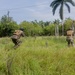 Three armed military or tactical personnel in camouflage uniforms and tactical gear are moving through a grassy field in what appears to be a tropical environment. The personnel are advancing in a tactical formation, with one individual in a crouched running posture suggesting a training exercise or field operation. The setting features dense tropical vegetation including palm trees and broad-leafed shrubs in the background.
