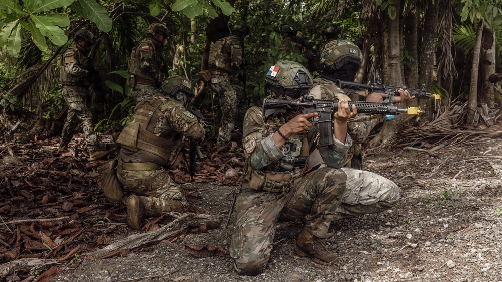 A group of military personnel in camouflage uniforms conduct what appears to be a jungle training exercise. The foreground soldier wears a helmet with a Panamanian flag patch and aims a rifle with a yellow training muzzle device, indicating a simulated exercise rather than live combat. Multiple soldiers are positioned in tactical stances within a dense tropical forest environment.