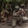 PANAMAX-26: V1/6, Partner forces conduct simulated air raid A group of military personnel in camouflage uniforms conduct what appears to be a jungle training exercise. The foreground soldier wears a helmet with a Panamanian flag patch and aims a rifle with a yellow training muzzle device, indicating a simulated exercise rather than live combat. Multiple soldiers are positioned in tactical stances within a dense tropical forest environment.