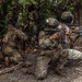 A group of military personnel in camouflage uniforms conduct what appears to be a jungle training exercise. The foreground soldier wears a helmet with a Panamanian flag patch and aims a rifle with a yellow training muzzle device, indicating a simulated exercise rather than live combat. Multiple soldiers are positioned in tactical stances within a dense tropical forest environment.
