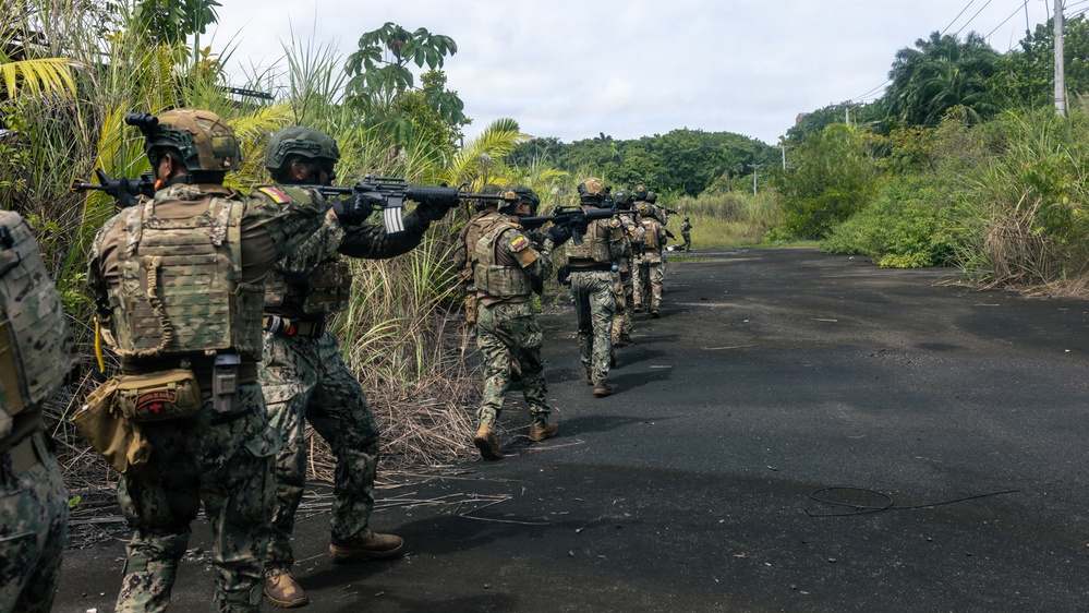 A column of military personnel in camouflage uniforms advances in tactical formation along a dark asphalt road flanked by dense tropical vegetation. The soldiers are armed and moving in a disciplined line with weapons raised in a ready or aimed position. At least one soldier displays a flag patch on their shoulder consistent with Ecuador's national colors.