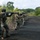 PANAMAX-26: V1/6, Partner forces conduct simulated air raid A column of military personnel in camouflage uniforms advances in tactical formation along a dark asphalt road flanked by dense tropical vegetation. The soldiers are armed and moving in a disciplined line with weapons raised in a ready or aimed position. At least one soldier displays a flag patch on their shoulder consistent with Ecuador's national colors.