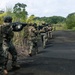 A column of military personnel in camouflage uniforms advances in tactical formation along a dark asphalt road flanked by dense tropical vegetation. The soldiers are armed and moving in a disciplined line with weapons raised in a ready or aimed position. At least one soldier displays a flag patch on their shoulder consistent with Ecuador's national colors.